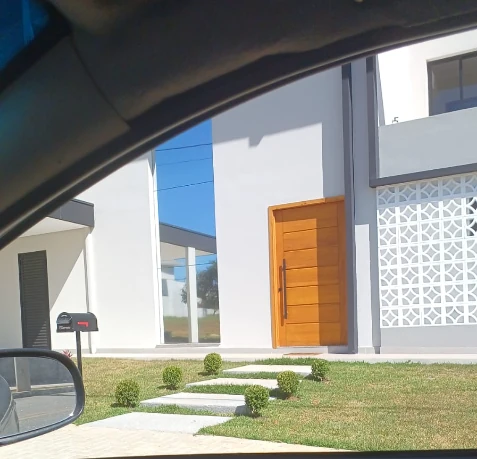 Fachada de casa com porta de madeira e jardim, fotografada de dentro de um veículo.