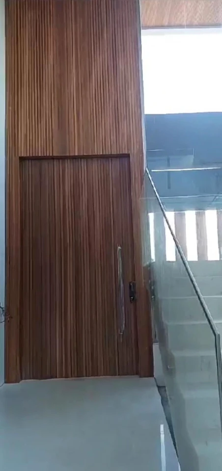 Porta de madeira ripada com bandeira superior e puxador vertical, ao lado de uma escada com guarda-corpo de vidro.
