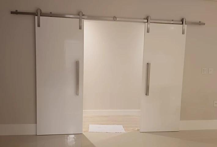 Porta branca de correr com puxador vertical e roldanas no trilho superior.