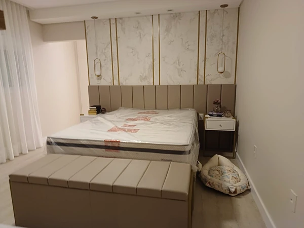 Cama com colchão embalado, cabeceira bege e banco estofado diante de painel claro com detalhes dourados.